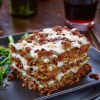 A slice of Pretto's Pasta "The Queen Lasagna" on a dark plate, showcasing multiple layers of delicate spinach pasta, creamy béchamel sauce, and rich meat Bolognese topped with melted cheese.